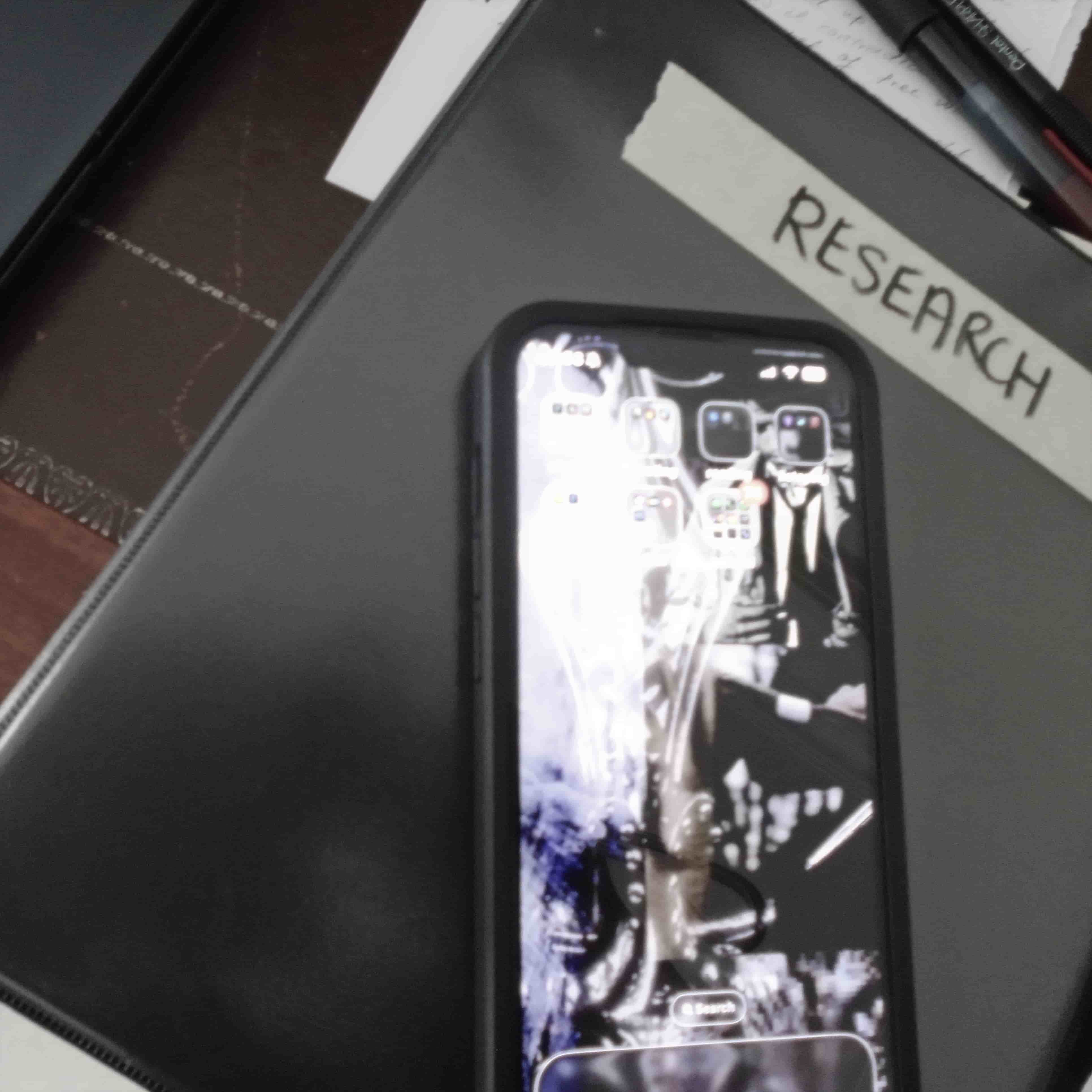 a phone illuminated on its home screen on top of a black binder with a masking tape label saying 'RESEARCH'