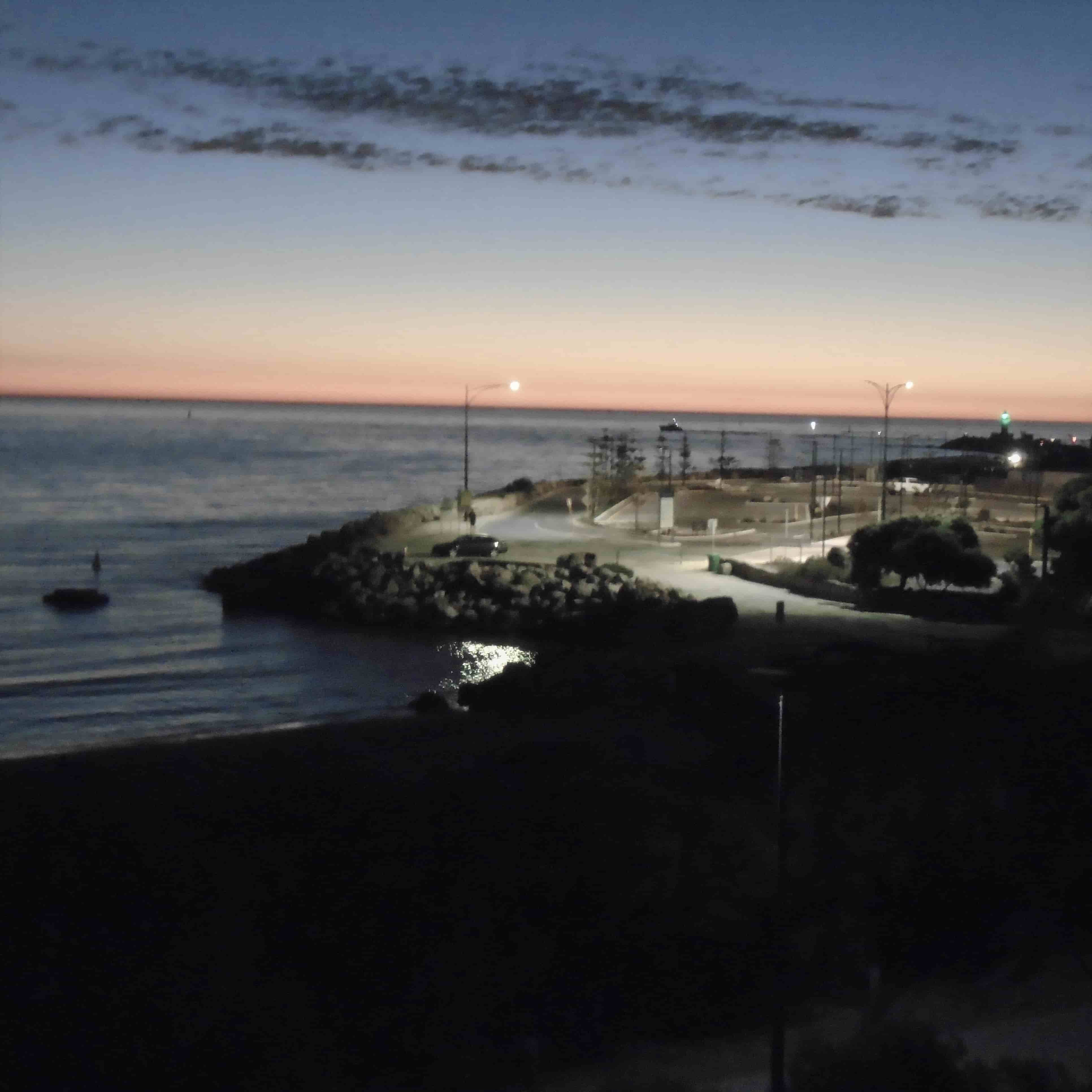 orange sunset on the horizon. beneath it, a rockly shoreline with roads and buildings is lit by streetlamps as it rests besides a dark blue sea